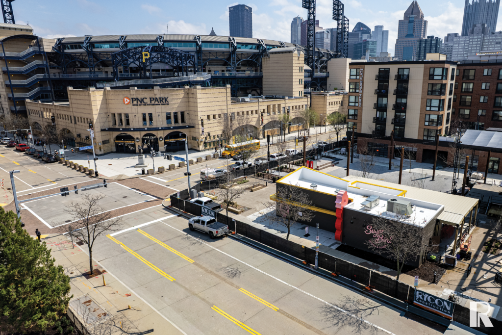 An elevated view of PNC Park in Pittsburgh with a nearby construction site and a “Sugar & Smoke” restaurant storefront in the foreground.