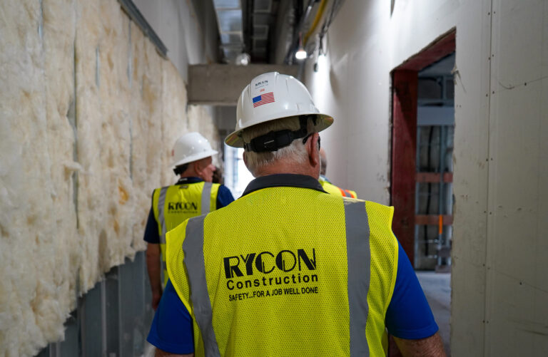 Three construction workers in hard hats and high-visibility safety vests walking down a hallway under construction.