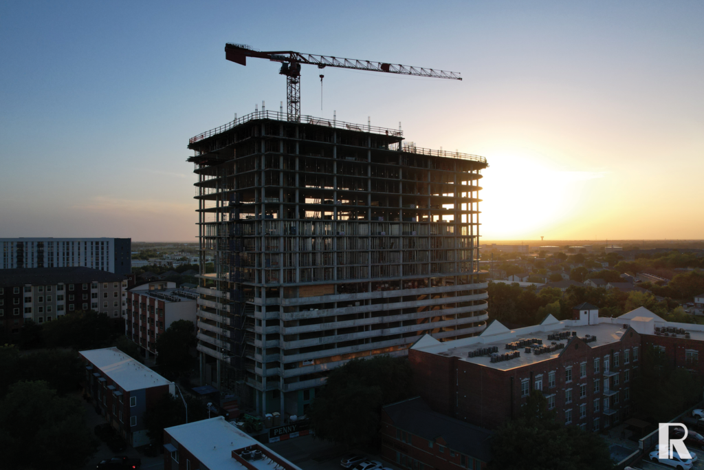 An aerial view of a multi-story building under construction with a tower crane on top, silhouetted against a setting sun.