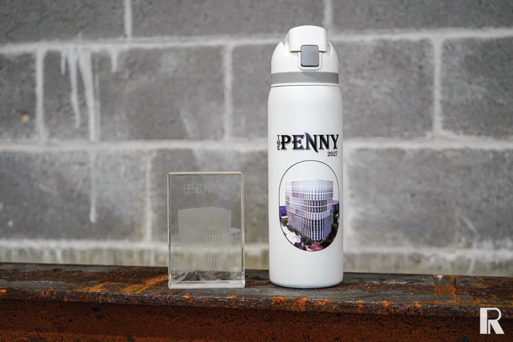 A white water bottle labeled “The Penny 2027” with an image of a modern building, sitting next to a glass block etched with a 3D architectural rendering of the same building.