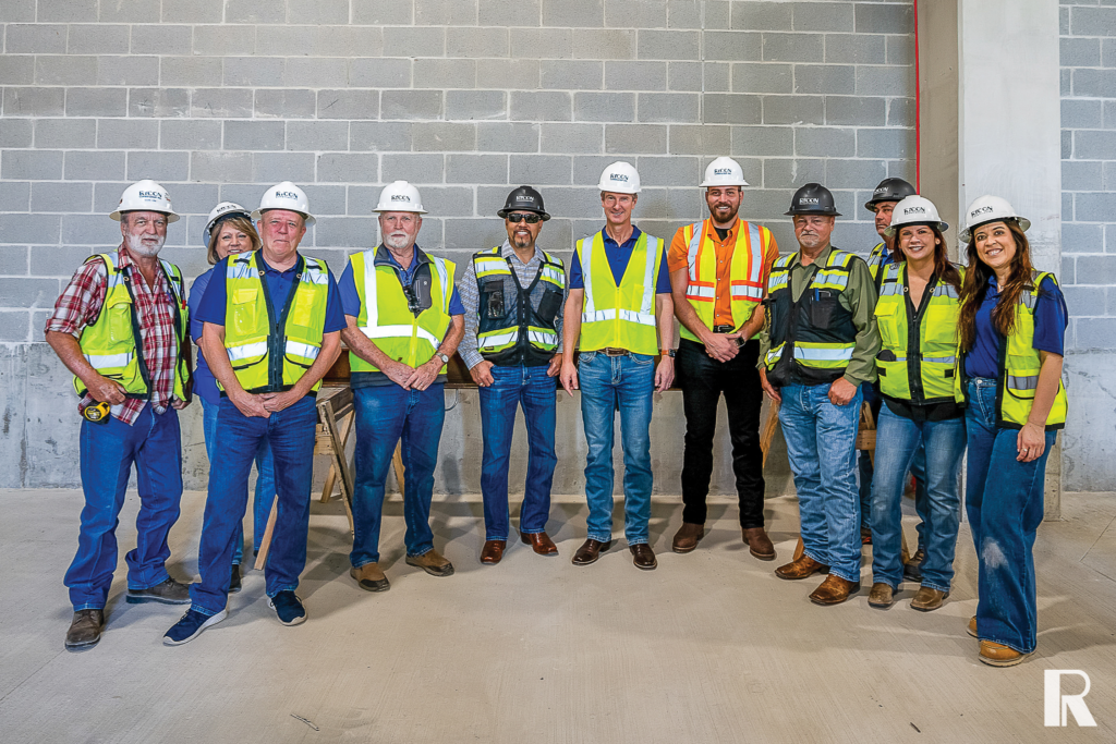 A group of eleven construction workers wearing safety vests and hard hats stand together in front of a concrete block wall.