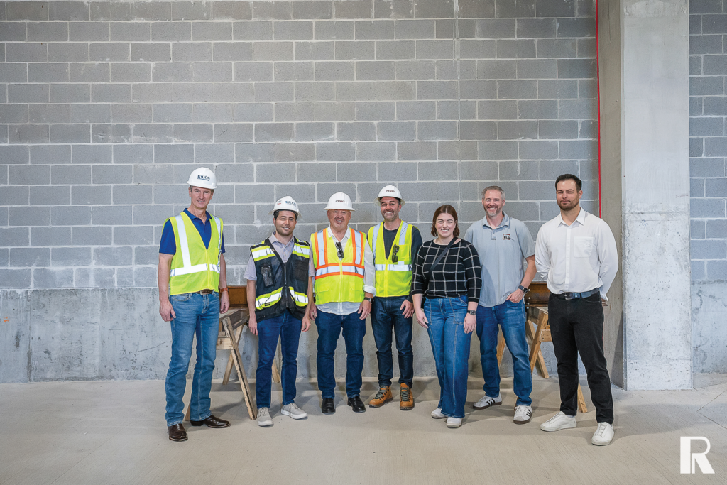 Seven people stand in a line against a gray concrete block wall in a construction site; most are wearing white hard hats and high-visibility vests.