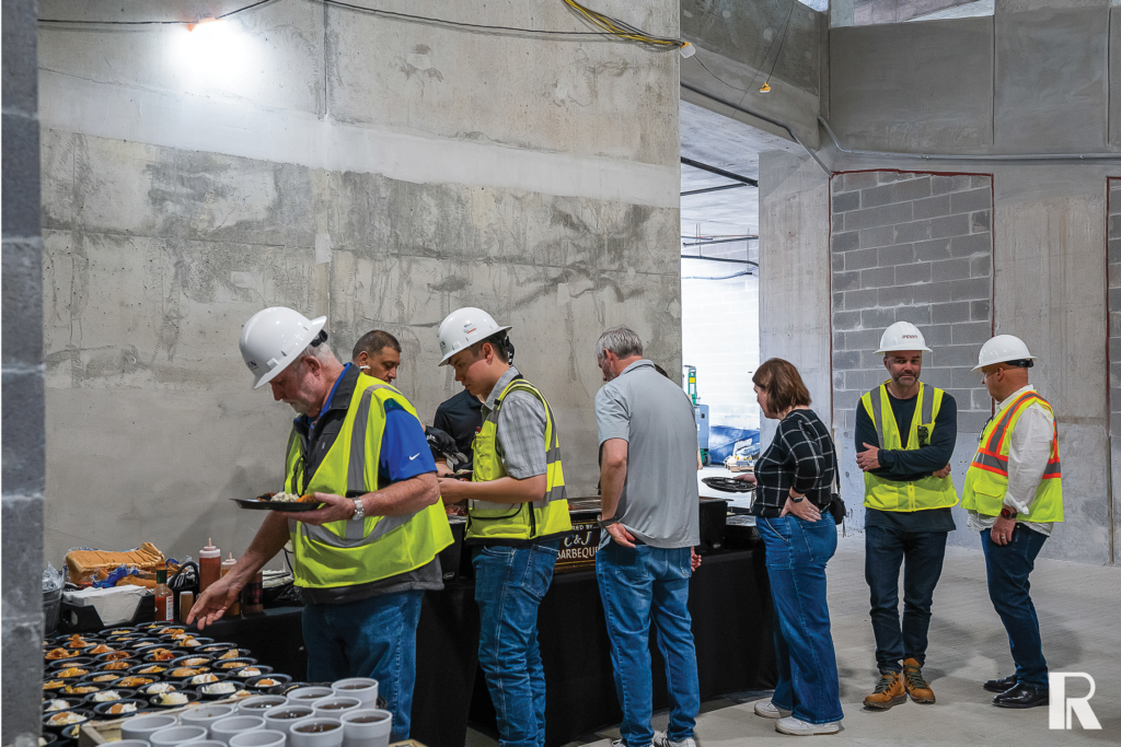 People in hard hats and high-visibility vests serve themselves food at a buffet line in an unfinished concrete building.