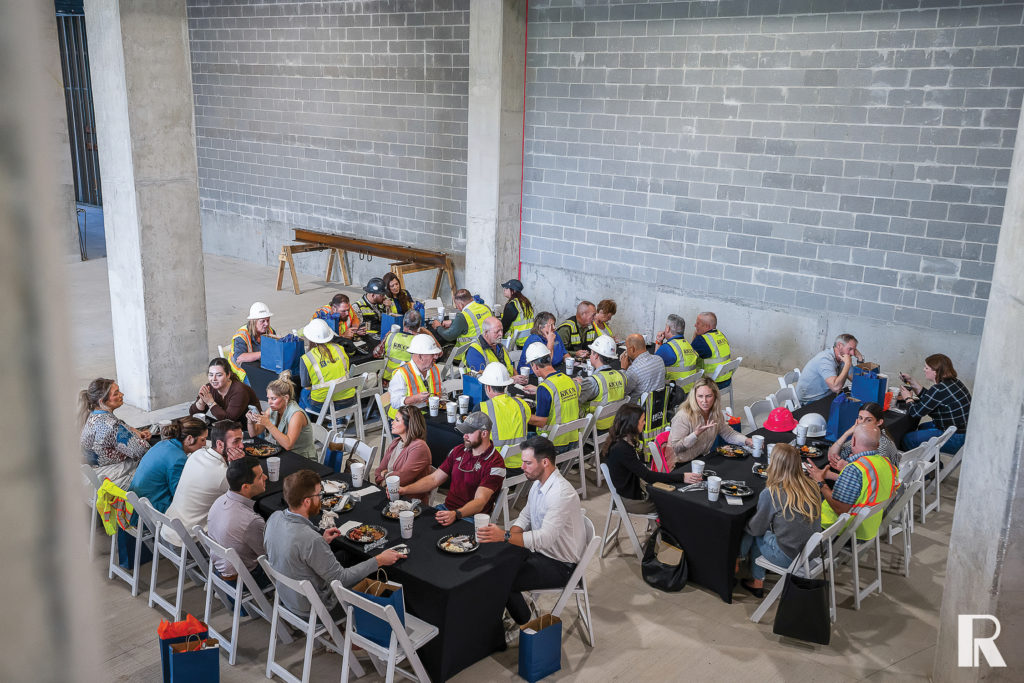 A group of people wearing safety vests and hard hats eat lunch together at tables in an unfinished construction space.