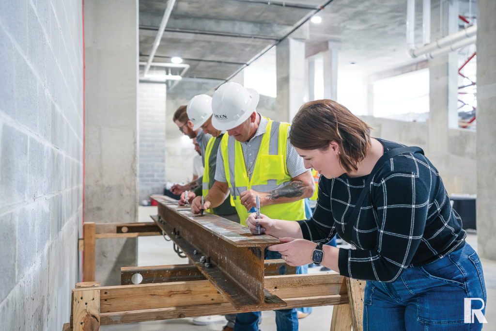 Four people in hard hats and high-visibility vests mark measurements on a steel beam at a construction site.