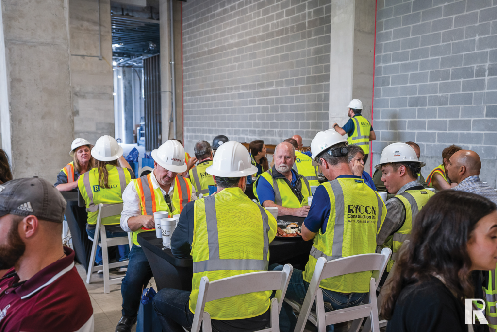 Construction workers wearing hard hats and high-visibility vests sit at tables eating lunch inside an unfinished building.