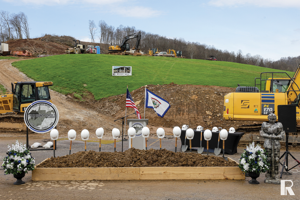 Groundbreaking ceremony for Tyler Consolidated Schools featuring a row of hard hats and shovels, an American flag, the West Virginia state flag, and construction equipment on a hillside.