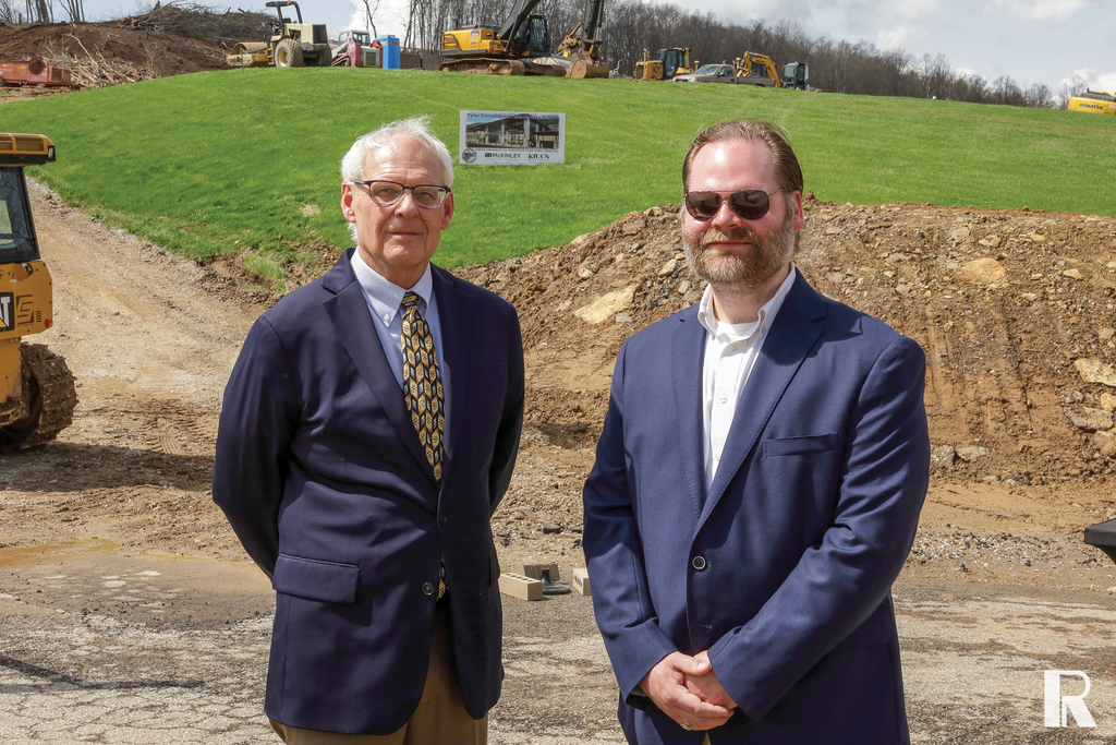 Two men in suits stand in front of a construction site with heavy machinery and a project sign in the background.