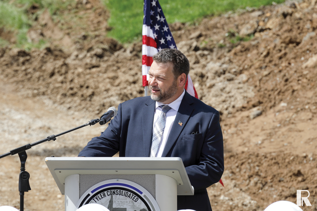 A man in a suit and tie speaks at a podium with an American flag in the background.