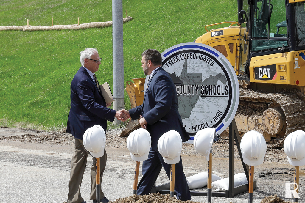 Two men in suits shake hands in front of a yellow bulldozer and a sign for Tyler Consolidated County Schools at a construction site with hard hats mounted on stakes.