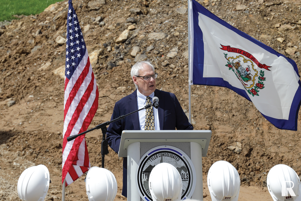 A man in a suit and tie speaks at a podium marked “Tyler Consolidated Schools,” flanked by the U.S. and West Virginia state flags with hard hats in the foreground.