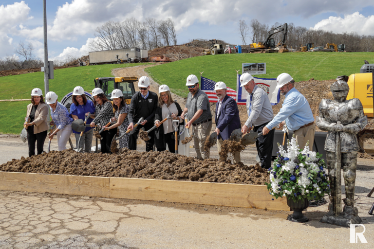 A group of people in hard hats participate in a ceremonial groundbreaking with shovels, standing before a pile of dirt and a decorative suit of armor.