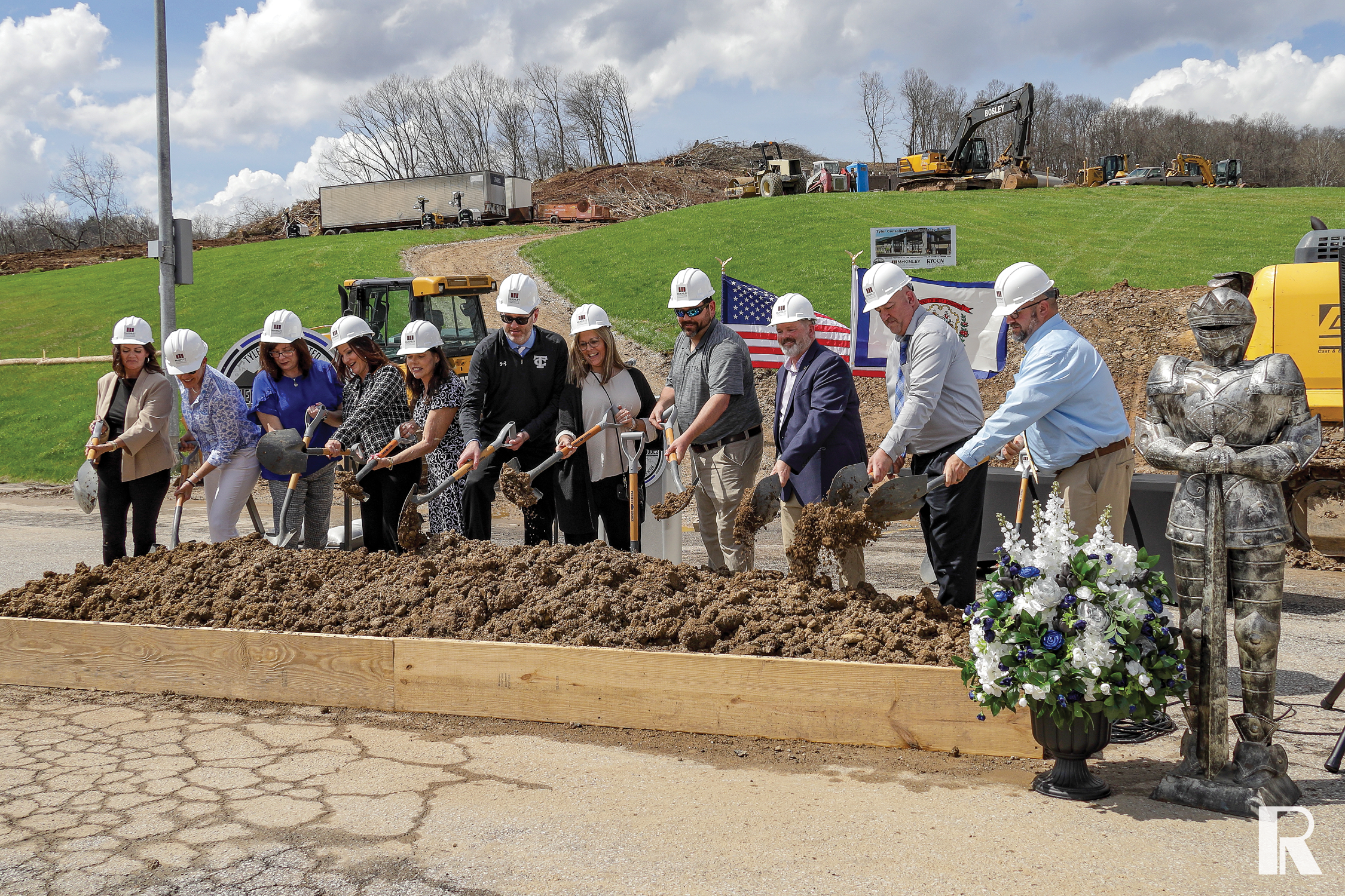 A group of people in hard hats participate in a ceremonial groundbreaking with shovels, standing before a pile of dirt and a decorative suit of armor.