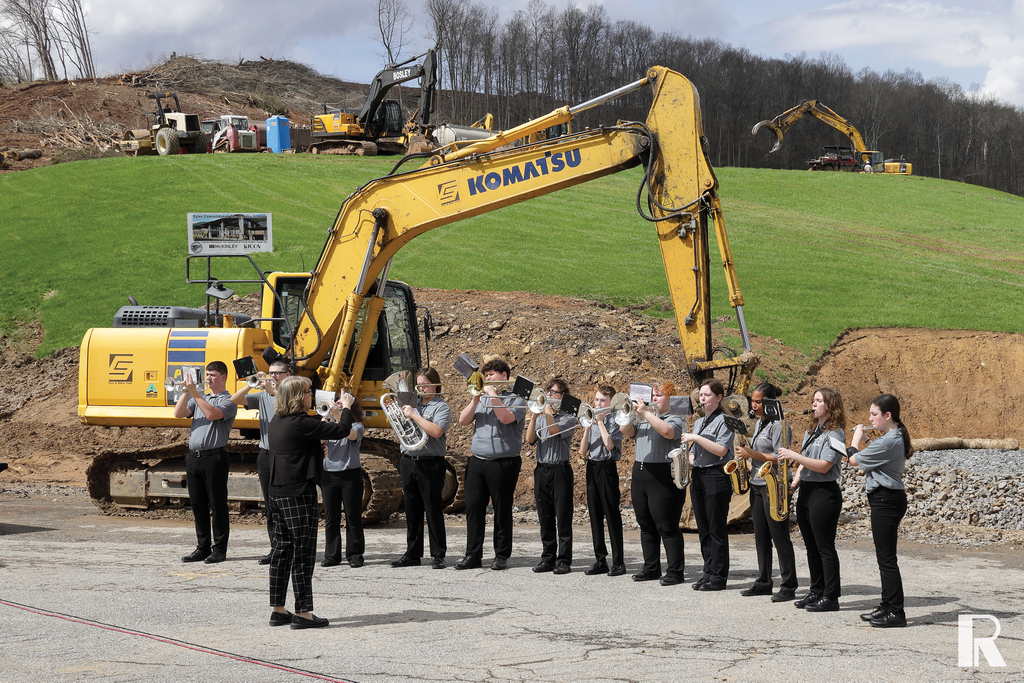 A high school marching band stands in a line playing brass instruments in front of a large yellow excavator at a construction site.