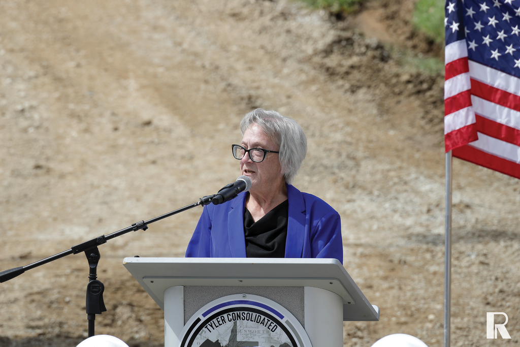 A woman with grey hair and glasses speaks at a podium with a Tyler Consolidated logo, with an American flag to her right.
