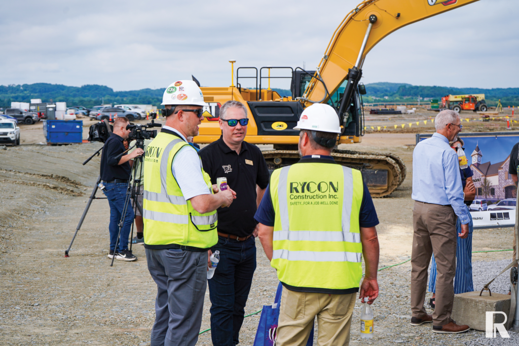Three men in construction safety gear and casual clothing stand on a dirt construction site, with a large yellow excavator in the background.