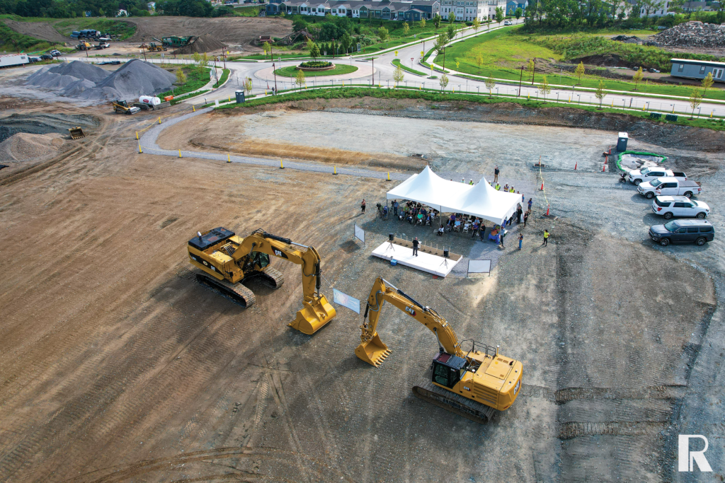 Aerial view of a ground-breaking ceremony with a white event tent and two yellow Caterpillar excavators on a construction site.