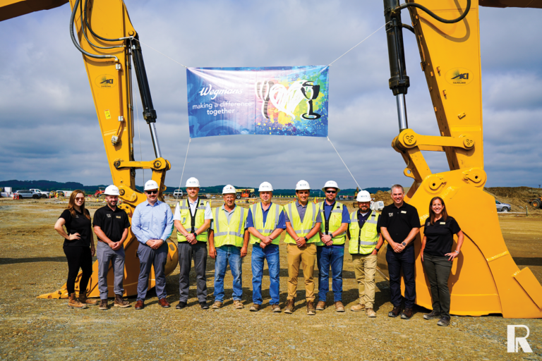 Eleven people in hard hats and work attire stand in a row beneath a banner reading “Wegmans making a difference together,” framed by the arms of two yellow construction excavators.