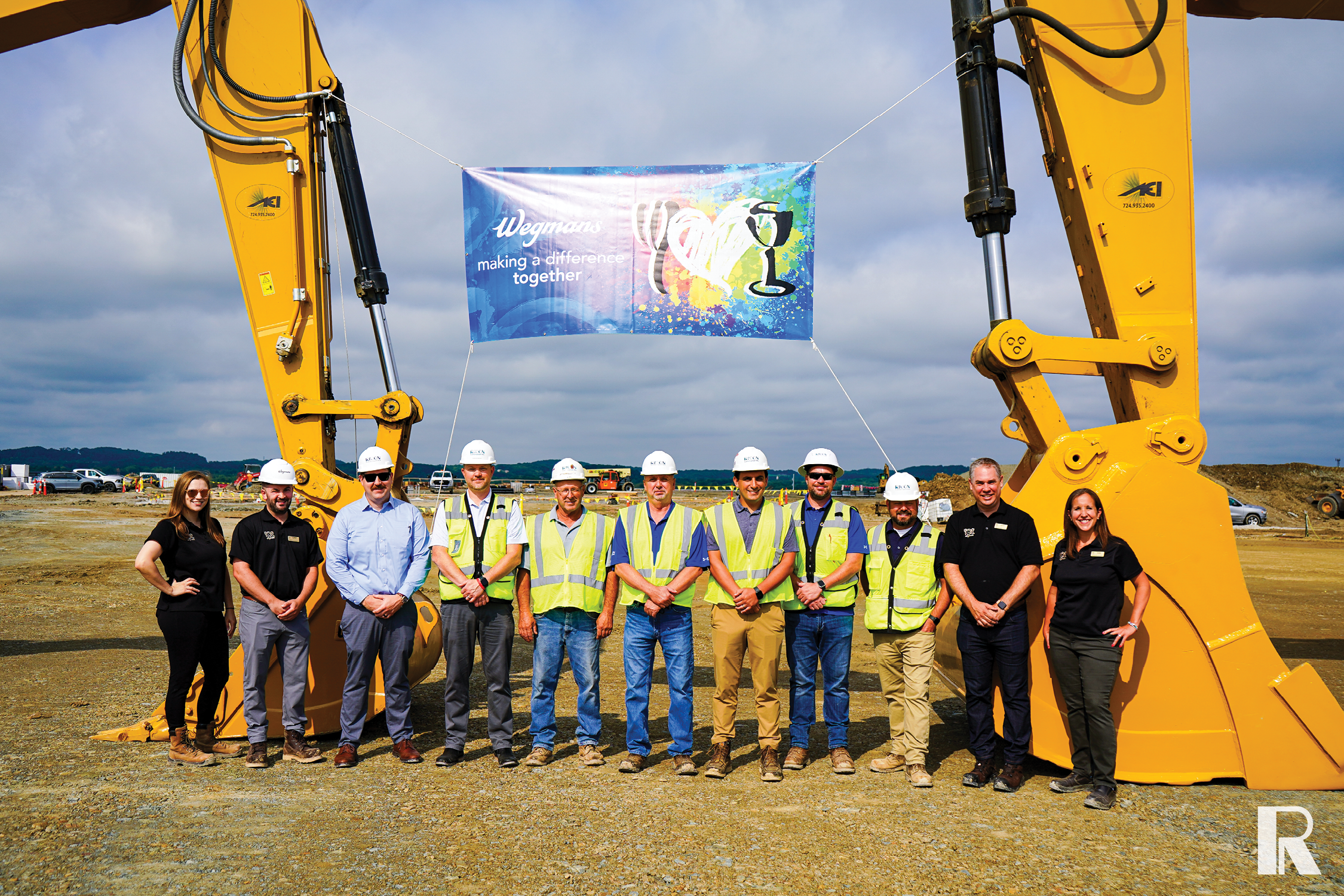 Eleven people in hard hats and work attire stand in a row beneath a banner reading “Wegmans making a difference together,” framed by the arms of two yellow construction excavators.