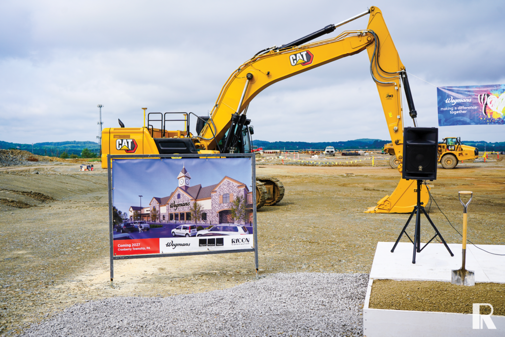 A construction site featuring a Caterpillar excavator and a sign displaying a rendering of a future Wegmans store, with a shovel and speaker in the foreground.