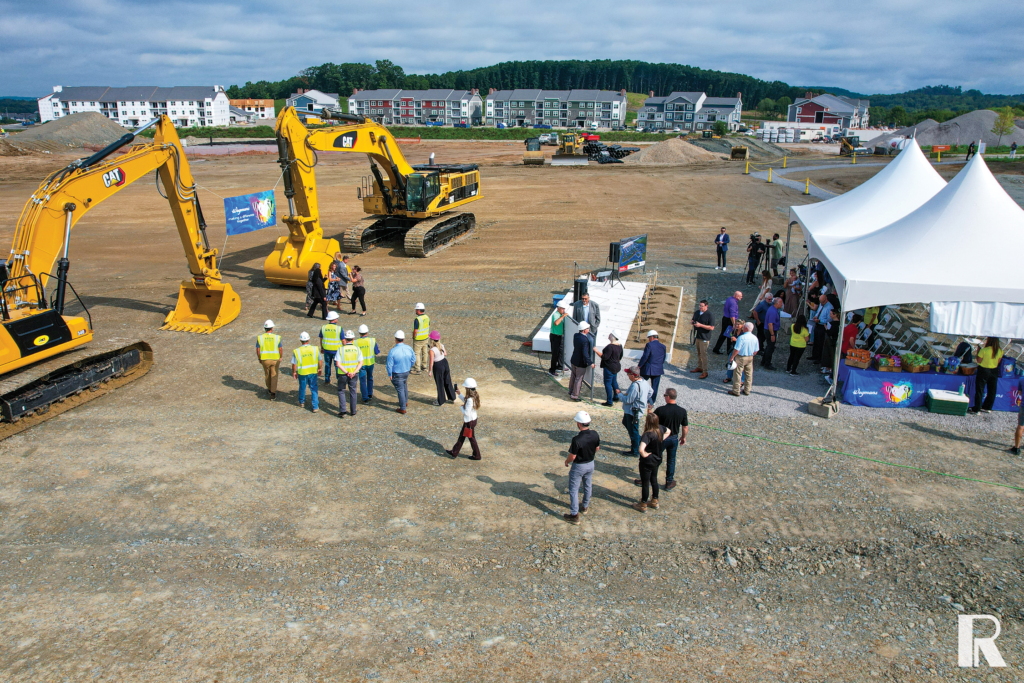 A group of people wearing hard hats and professional attire gather at a construction site with two large yellow excavators and a white event tent.