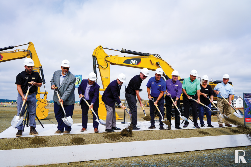 Ten people in hard hats participate in a ceremonial groundbreaking with shovels in front of yellow construction excavators.