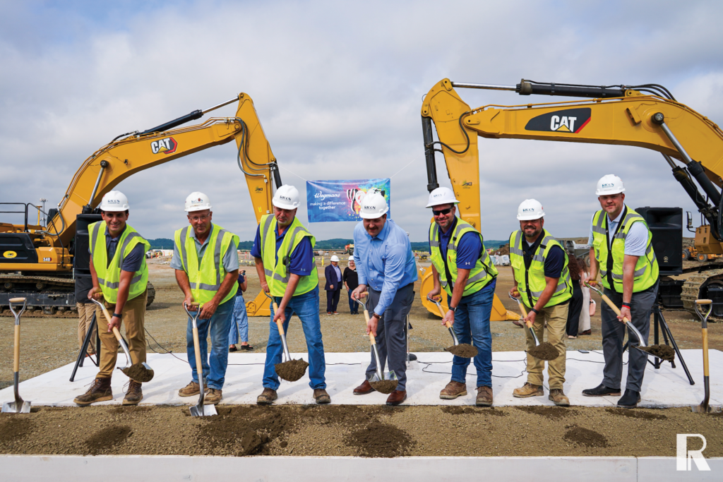 Seven people in hard hats and high-visibility vests hold shovels to break ground at a construction site in front of two yellow excavators and a Wegmans banner.