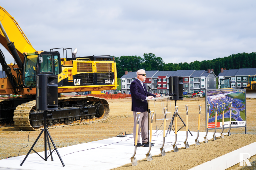 A man in a suit speaks at a podium during a ground-breaking ceremony at a construction site with a yellow excavator and a site plan sign nearby.