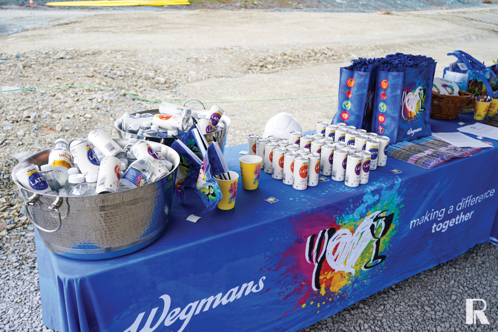 A Wegmans branded table with cans of latte in ice buckets, paper cups, and blue gift bags.