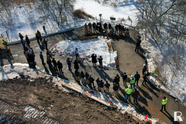 A group of people wearing hard hats perform a ceremonial groundbreaking with shovels at a snowy construction site, while spectators watch from the perimeter.