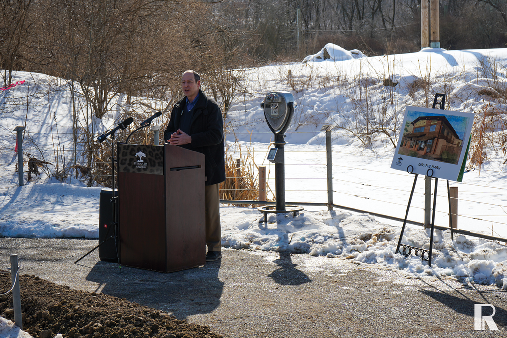 A man speaks at a podium outdoors in a snowy setting next to an easel displaying a rendering of a building labeled “Giraffe Barn.”