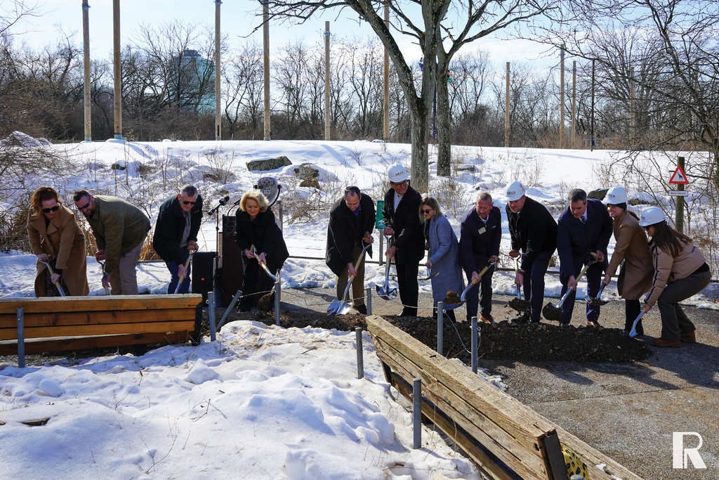 A group of people in business attire and hard hats participate in a ceremonial groundbreaking at a snow-covered park.
