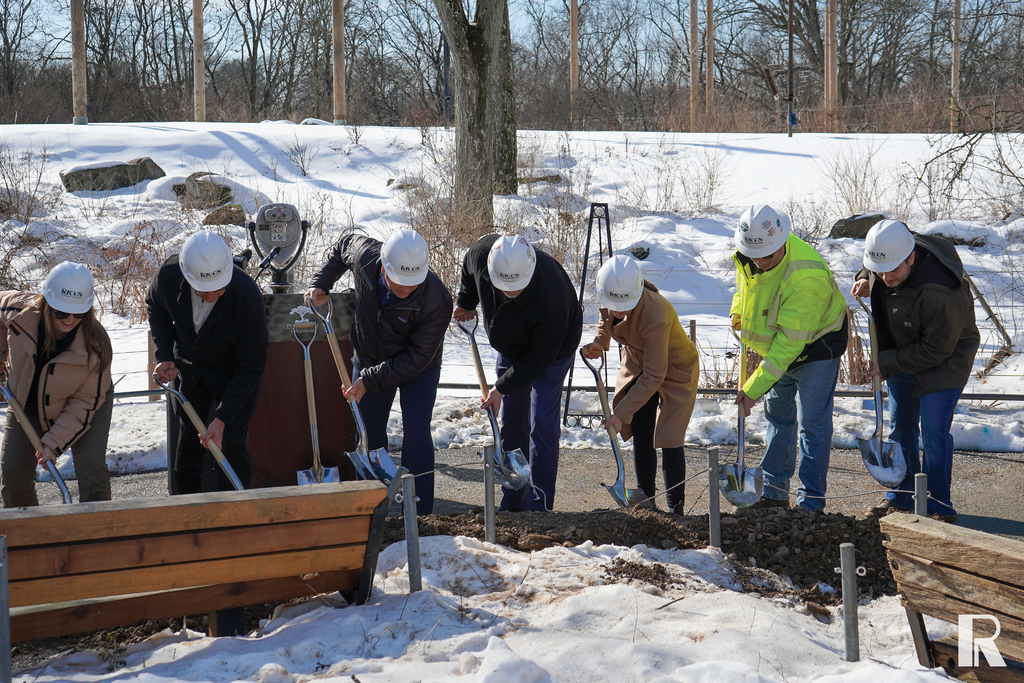 A group of people in hard hats participate in a ceremonial groundbreaking, using shovels to dig into a patch of dirt in a snowy park.