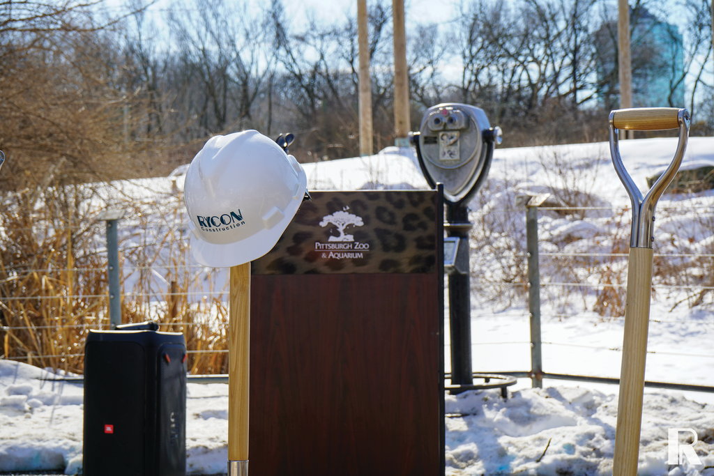 A white Rycon Construction hard hat and a shovel on display in front of a Pittsburgh Zoo & Aquarium podium in a snowy outdoor setting.