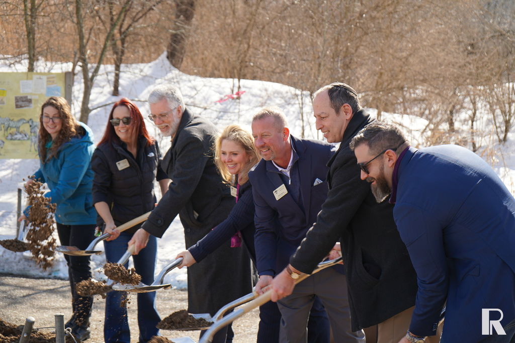 A group of people wearing name tags ceremonially shovel dirt during an outdoor groundbreaking event in a snowy setting.