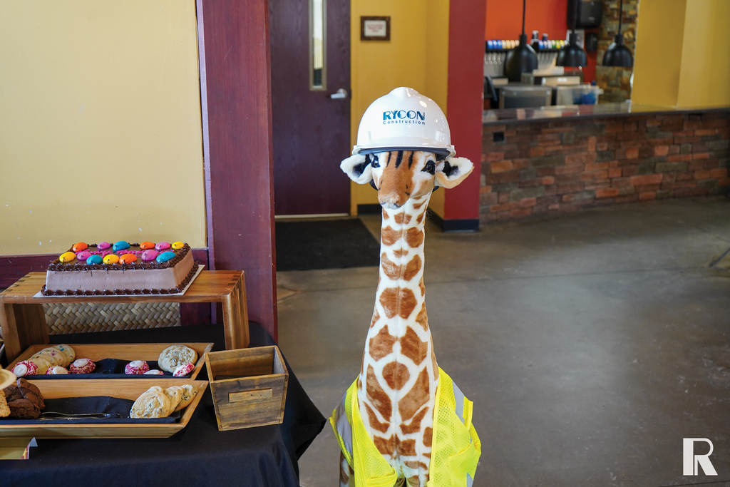 A plush giraffe toy wearing a Rycon Construction hard hat and a high-visibility safety vest, standing next to a table with trays of cookies and a cake.