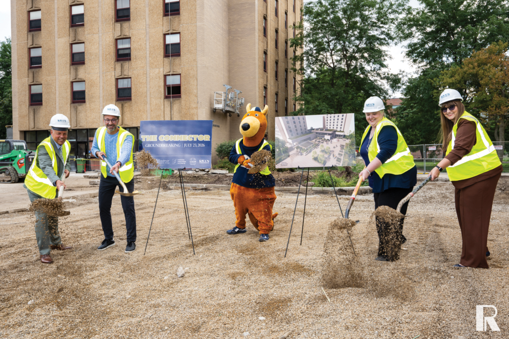 Five people, including a mascot in a bear costume, wearing hard hats and high-visibility vests participate in a ceremonial groundbreaking at a construction site.