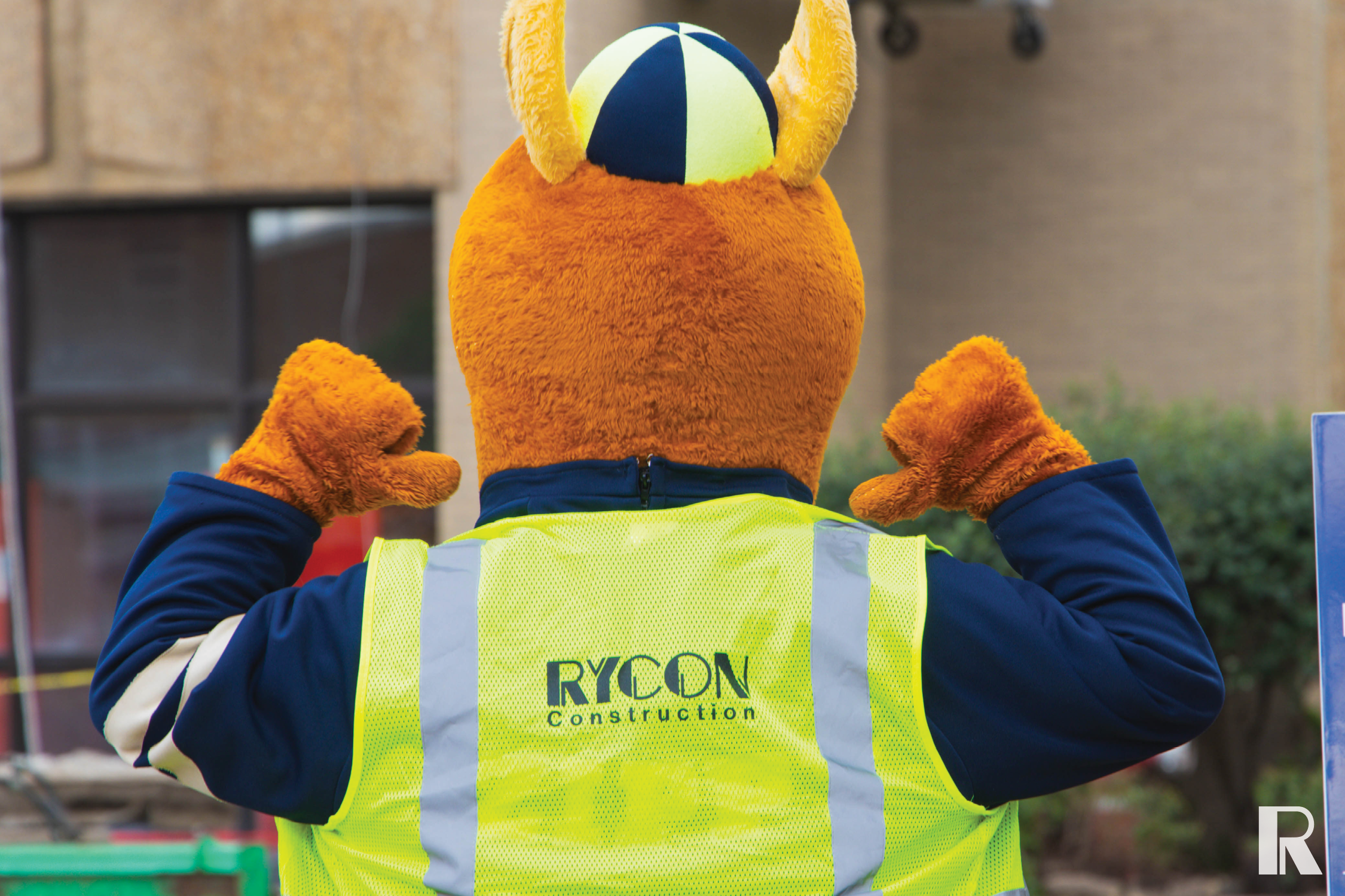 A person in an orange mascot costume wears a yellow high-visibility safety vest with the “Rycon Construction” logo on the back.