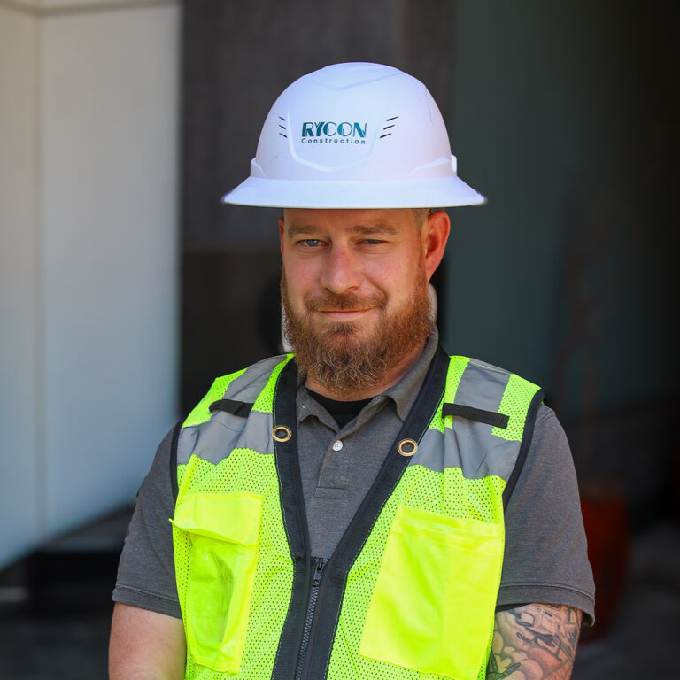 A man with a beard wearing a white Rycon Construction hard hat and a high-visibility safety vest.