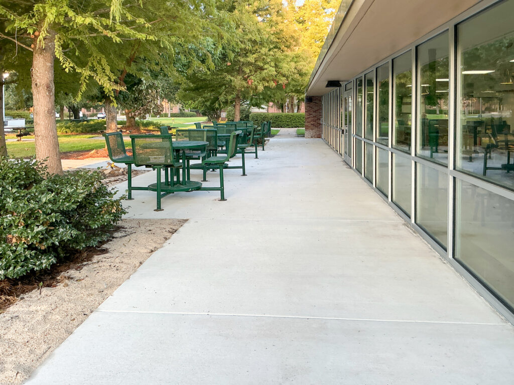 Concrete walkway with green metal outdoor tables and chairs outside a glass-walled building.