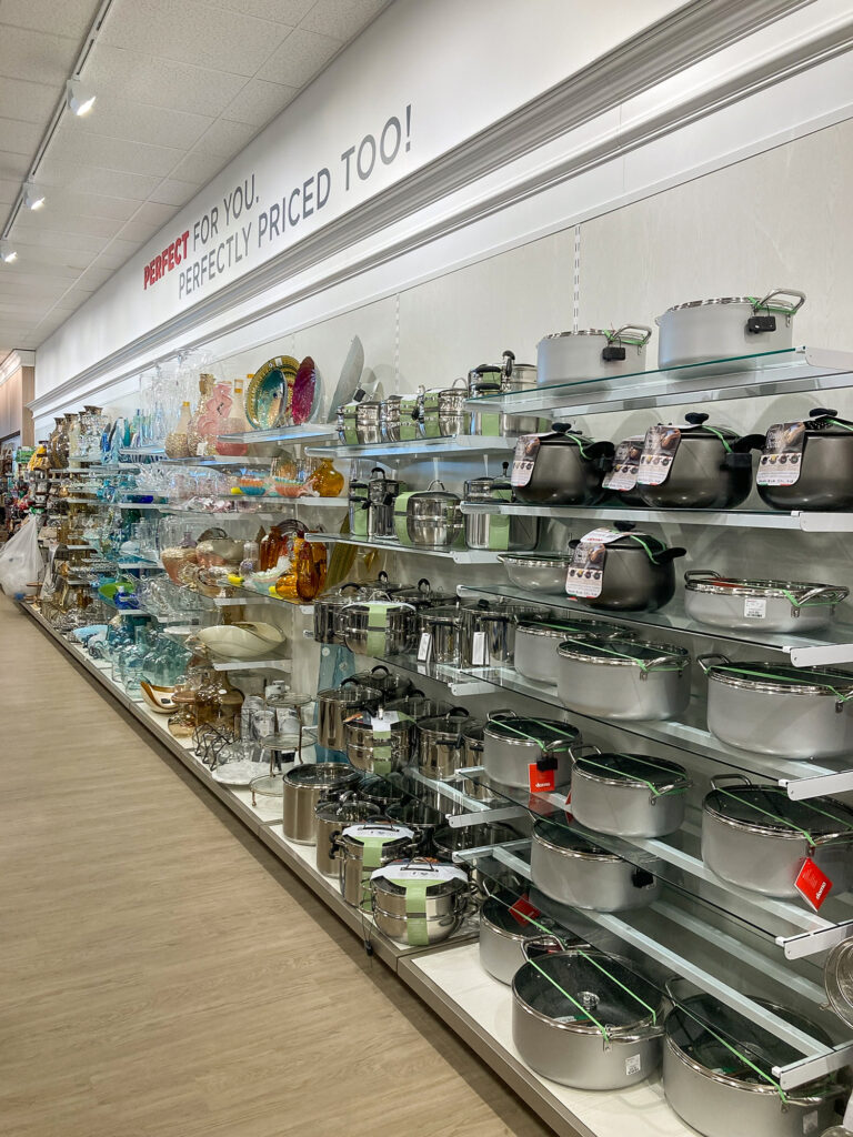 Store shelving display featuring decorative glassware on the left and a large selection of stainless steel pots and pans on the right.