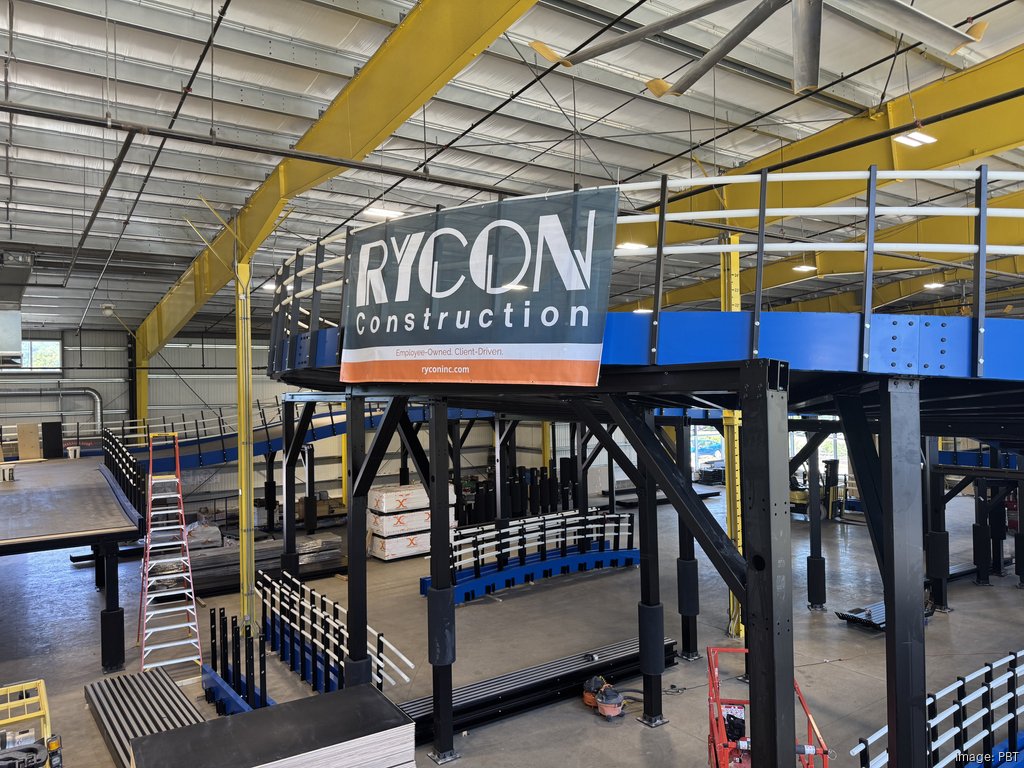 A banner for Rycon Construction hangs on the railing of a raised metal platform inside a large warehouse.