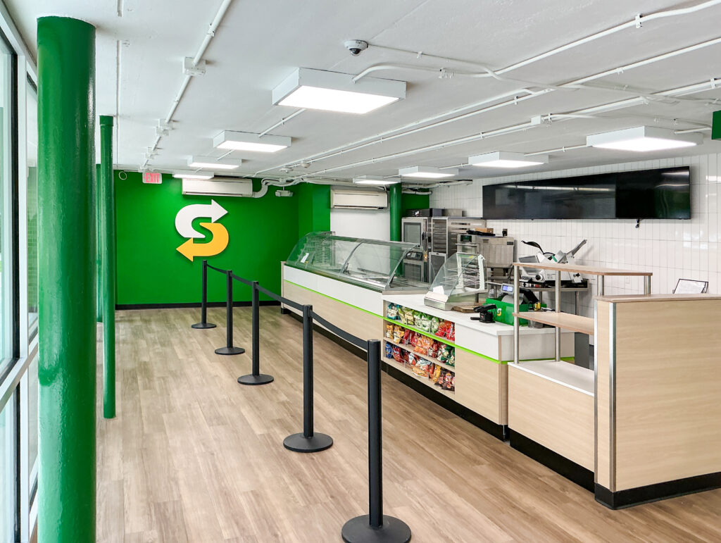 Interior of a Subway restaurant with a service counter, a display case of snacks, and stanchions for a customer queue.