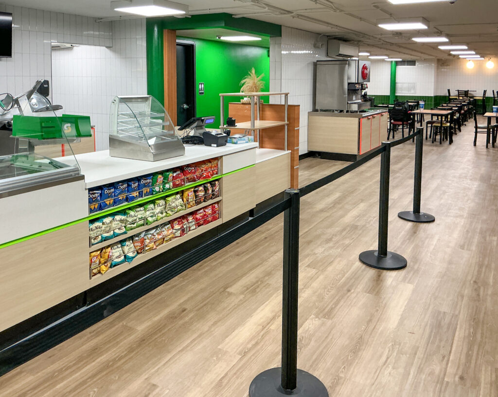 Interior of a Subway restaurant featuring a snack display under the service counter, a queue stanchion, and dining tables in the background.