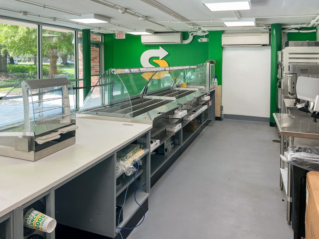Interior of an empty Subway restaurant featuring a stainless steel serving counter and a green accent wall with the brand logo.