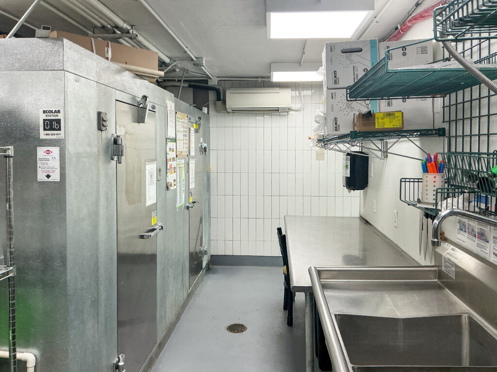 Commercial kitchen area with a large walk-in cooler, stainless steel work table, and utility sink.