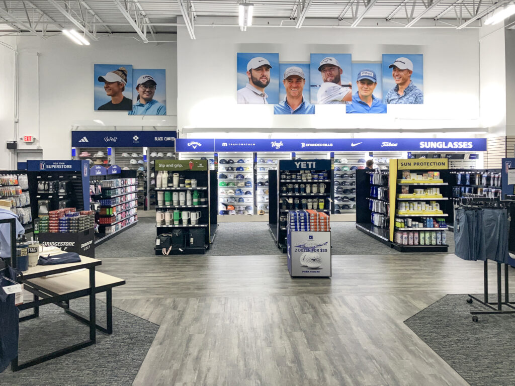 Interior view of a golf retail store with shelves of equipment, apparel, and accessories, featuring large portraits of professional golfers on the wall.