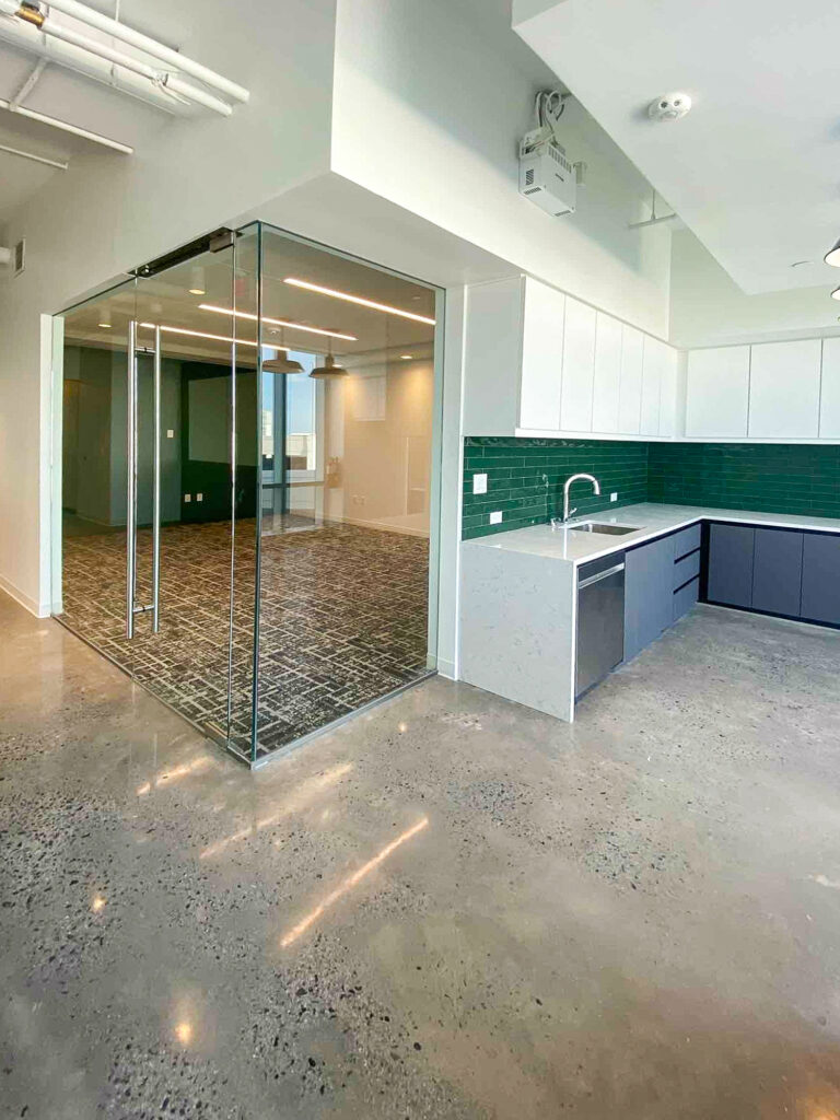 An office interior with a glass-walled conference room next to a kitchenette with white upper cabinets, dark lower cabinets, and a green tiled backsplash.