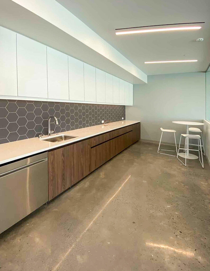 Modern office kitchenette with white upper cabinets, wood-tone base cabinets, a gray hexagonal tile backsplash, and a small round table with stools.