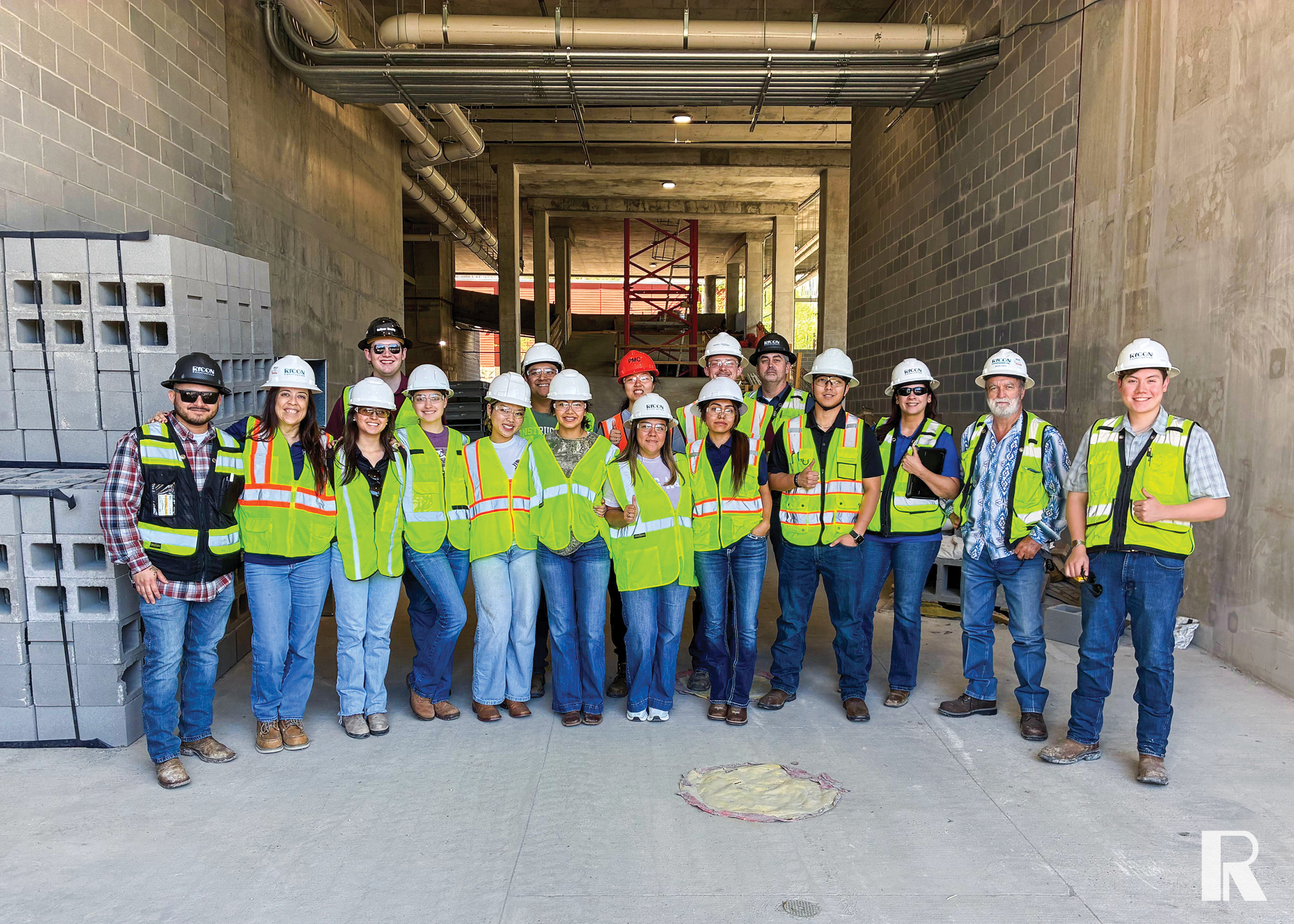 A group of construction workers in hard hats and high-visibility vests posing for a photo inside an unfinished concrete building.